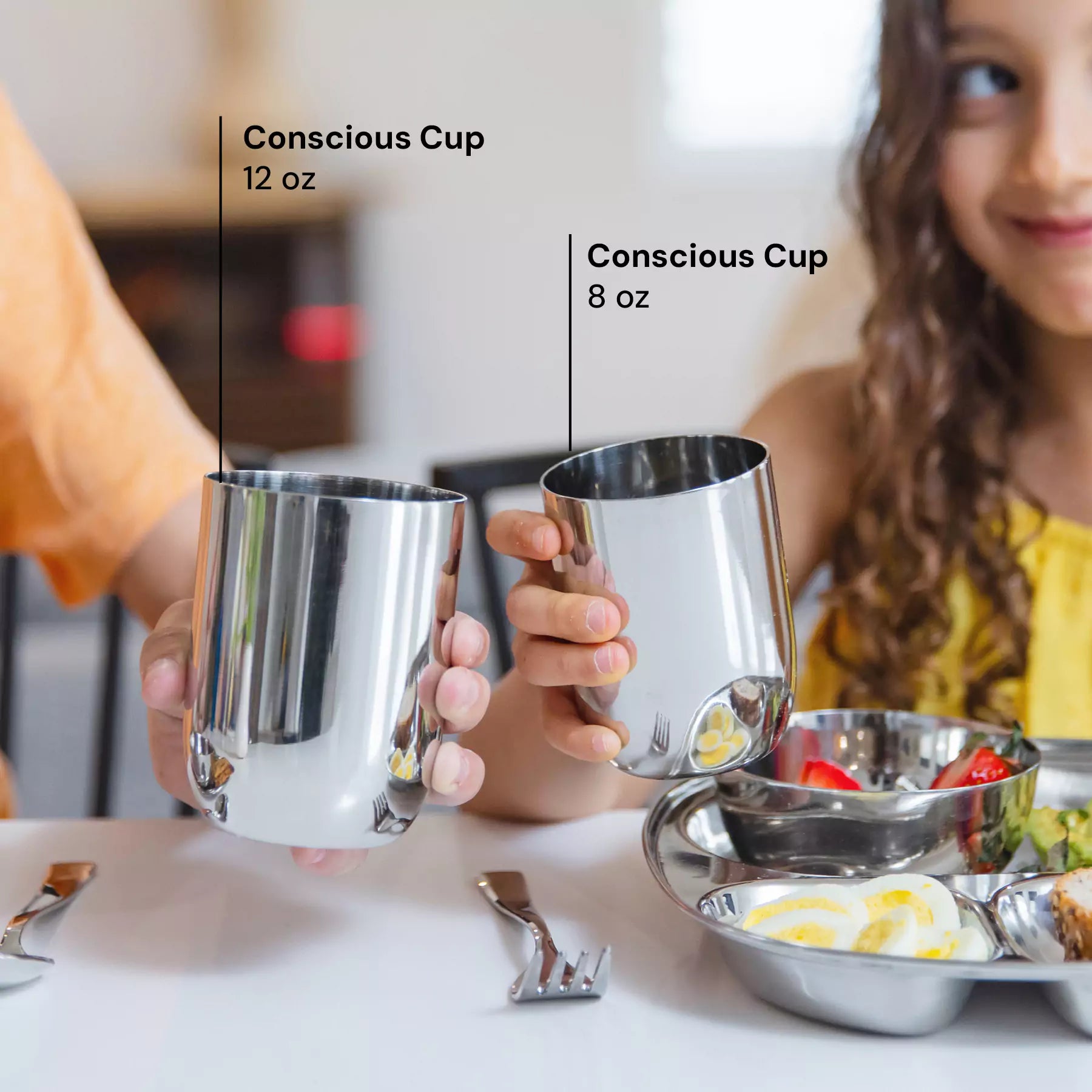 Two kids with Conscious Cups, on the left side the 12 ounce stainless steel shiny cup and on the right side an 8 ounce stainless steel cup reflecting the child's food. 