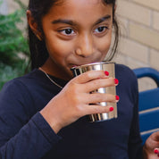 Young girl drinking from a stainless steel, shiny 10 ounce cup drinking water outdoors on a bench. The perfect size for kids, reduces waste, dishwasher safe, durable, and the healthiest cup for kids