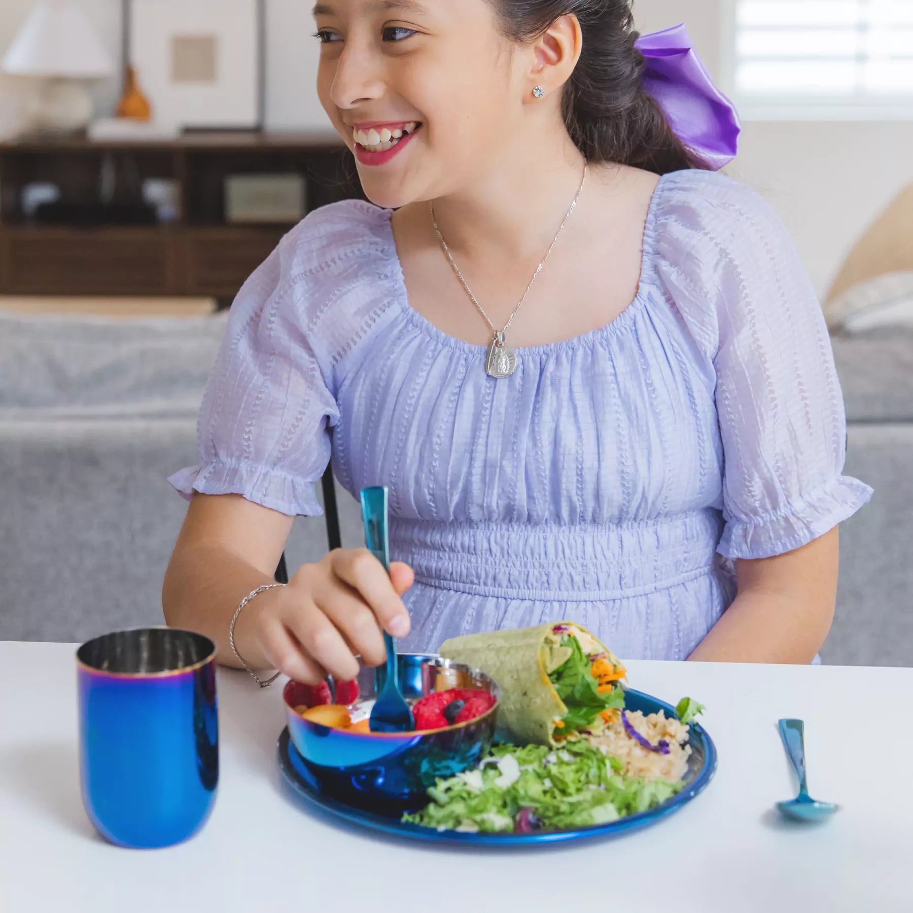 Girl (older kid, around 11 years old) in a light blue dress eating a meal at a table on Ahimsa's stainless steel Purposeful Portions Set, which includes plate, bowl, cup, fork, and spoon