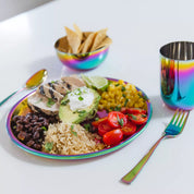 Stainless steel rainbow plate of food with rice, beans, avocados, corn, and tomatoes, accompanied by a rainbow-colored stainless steel 12 ounce cup, 14 ounce stainless steel bowl, fork and spoon on a white surface.