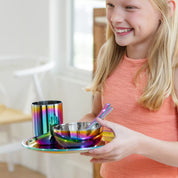 Child holding a rainbow-colored stainless steel dining set with a bowl, cup, plate, fork, and spoon